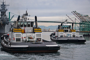 Two compact tugboats traveling side by side across a harbor, each with a white cabin, black hull, and yellow equipment mounted near the deck.