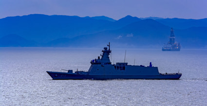 Silhouette of a modern naval ship moving across calm water, viewed from the side against a blue-toned seascape