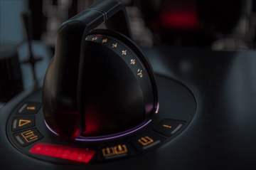 Close-up of two joystick-style control levers mounted on a dark console, each surrounded by illuminated circular indicators in red and purple tones. 