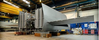 Large, angular metal ship structure inside an industrial workshop, likely a section of a vessel under construction.