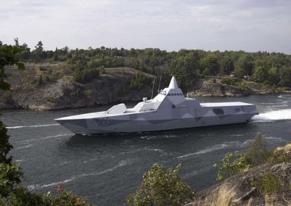 Grey stealth naval vessel with an angular, faceted design traveling at speed through a narrow coastal channel