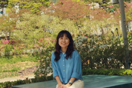 Person seated on a wooden bench in a landscaped park, facing the camera. The park features dense greenery, trees with sunlit leaves, and flowering shrubs in the background.