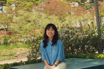 Person seated on a wooden bench in a landscaped park, facing the camera. The park features dense greenery, trees with sunlit leaves, and flowering shrubs in the background.