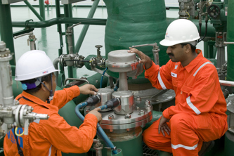 Two offshore workers wearing bright orange protective coveralls and white hard hats are performing maintenance on a large green industrial pipe system