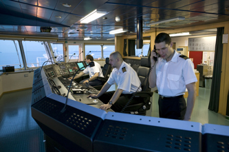 Interior of a ship’s bridge with three crew members operating navigation and control consoles.