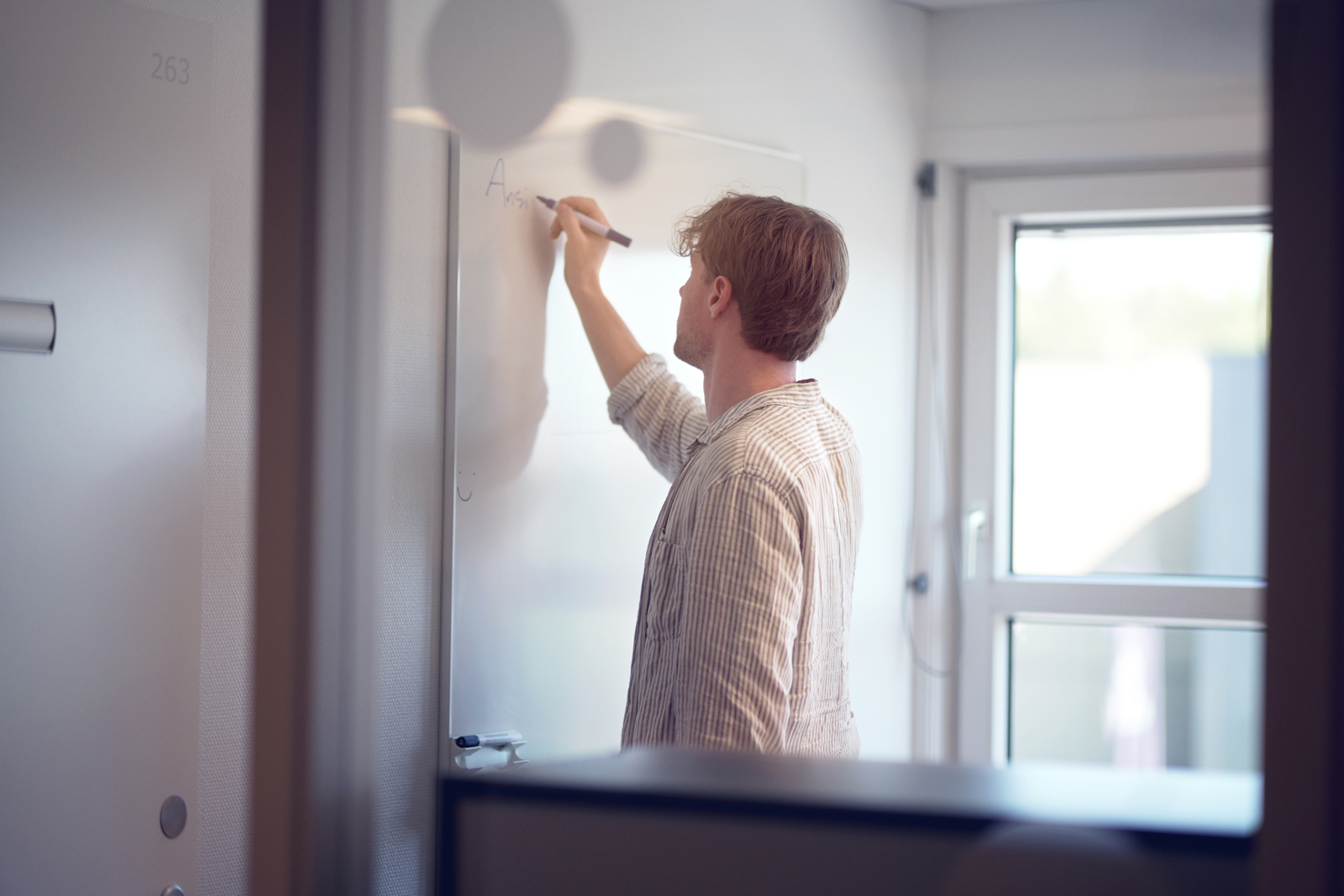 Person standing in a small office or meeting room, writing on a whiteboard mounted on the wall.