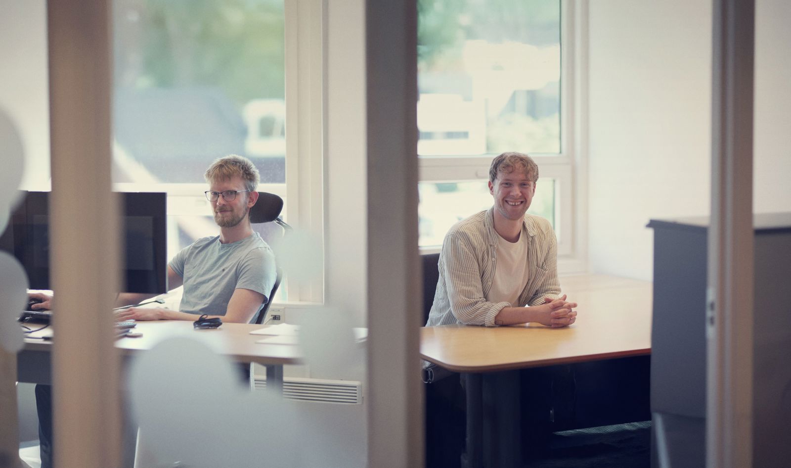 Two people seated at desks in a bright office, viewed through a glass partition. One person sits at a workstation with computer monitors on the left, while another sits at a desk on the right, facing the camera.