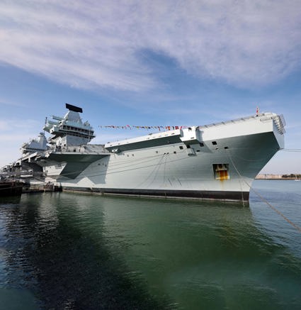 Large grey aircraft carrier docked at a harbor, viewed from a low angle near the bow.