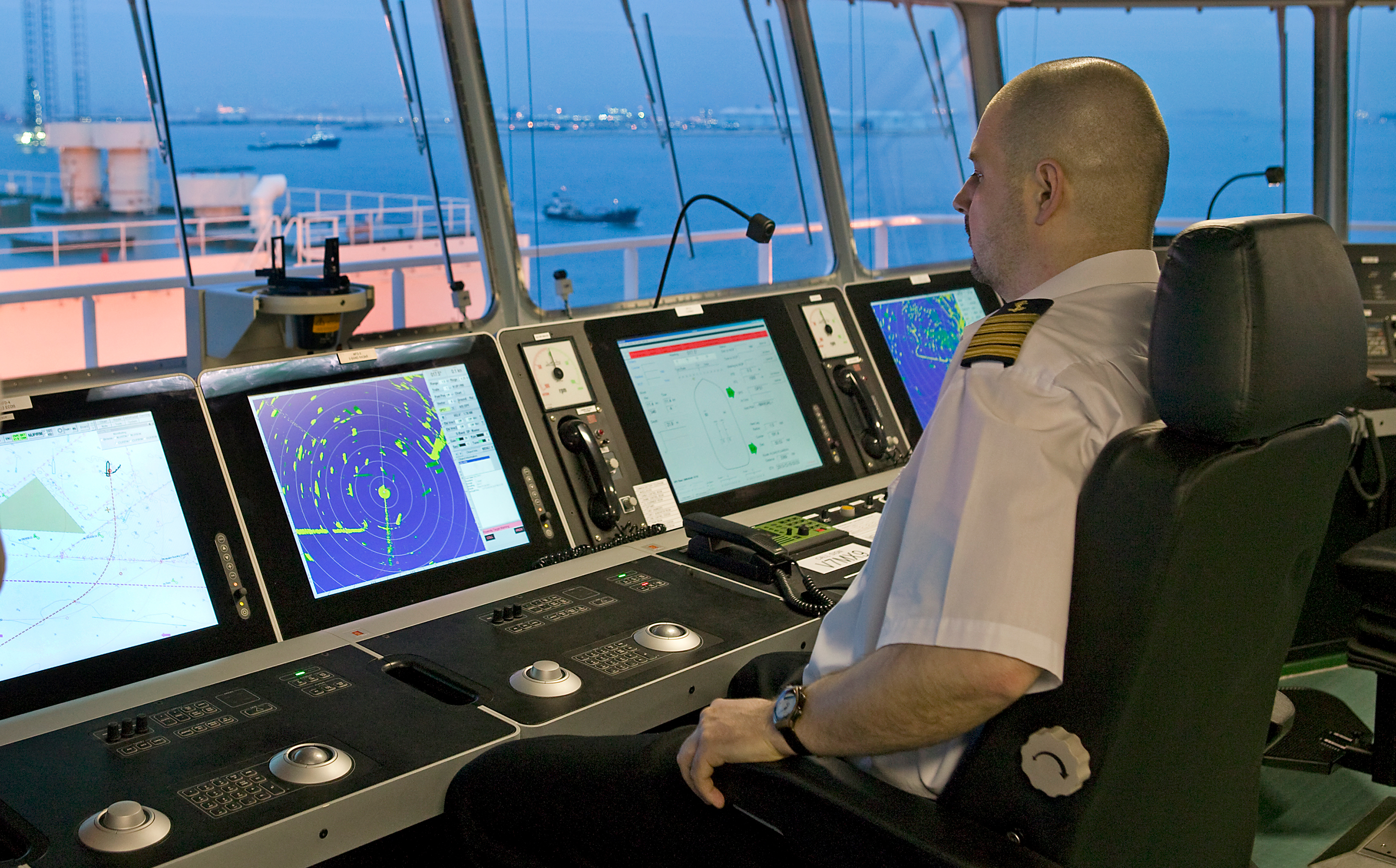 Ship bridge control room with a crew member seated at a console monitoring multiple navigation and radar displays, including charts and vessel traffic. 