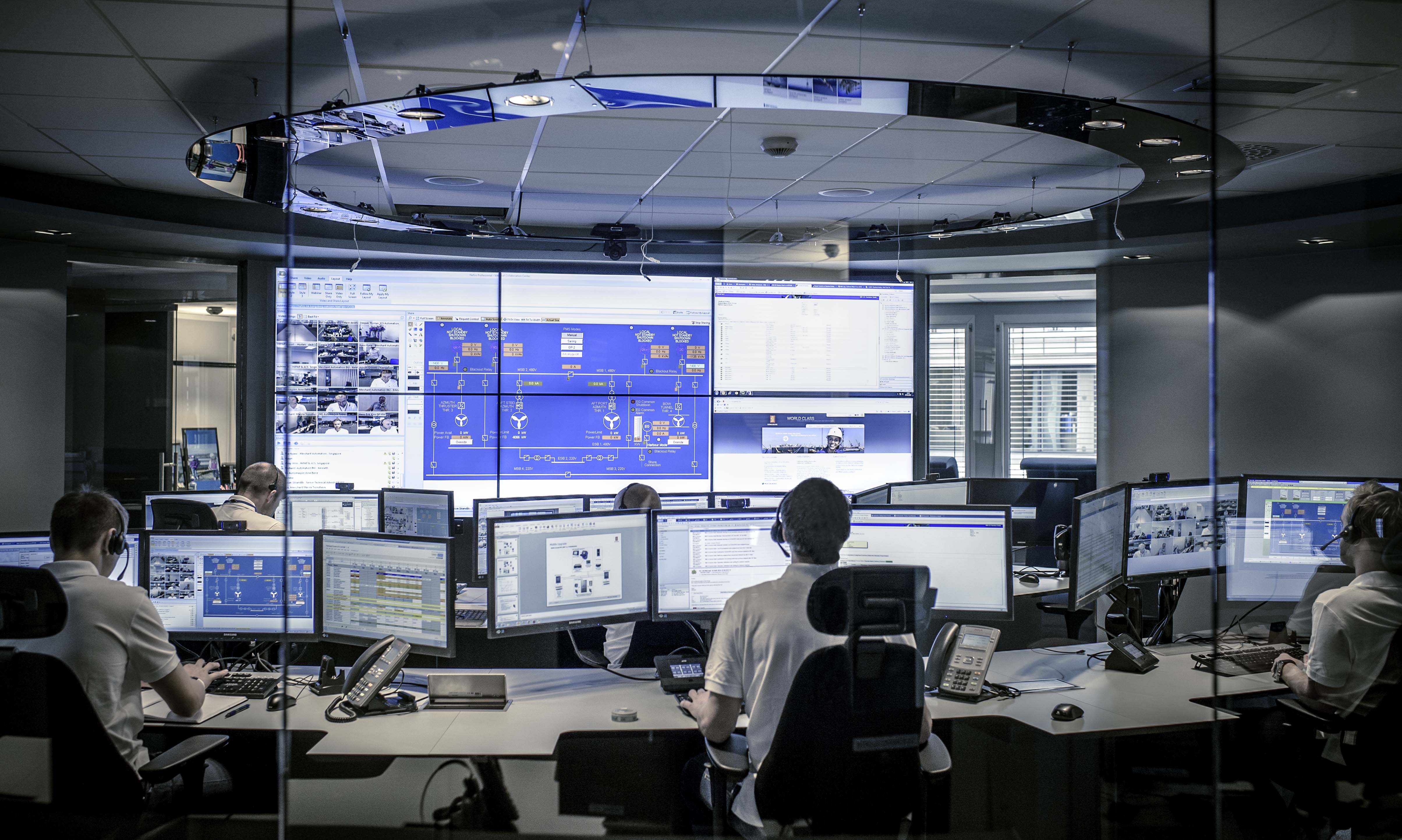 Wide view of a Kongsberg Maritime customer support centre, with several employees handling customer enquiries on phone and by email via large dual computer desktops and a large system of wall-mounted screens showing system overview and videoconference call