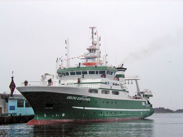 Green and white research vessel labeled “CELTIC EXPLORER” docked beside a pier in calm water. The ship features multiple decks with antennas, radar equipment, and communication masts on top.