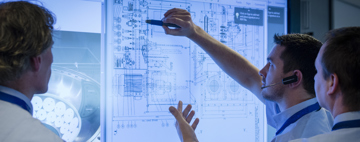Engineers reviewing a detailed technical drawing on a large digital display, with one person pointing to key design elements during a collaborative engineering discussion
